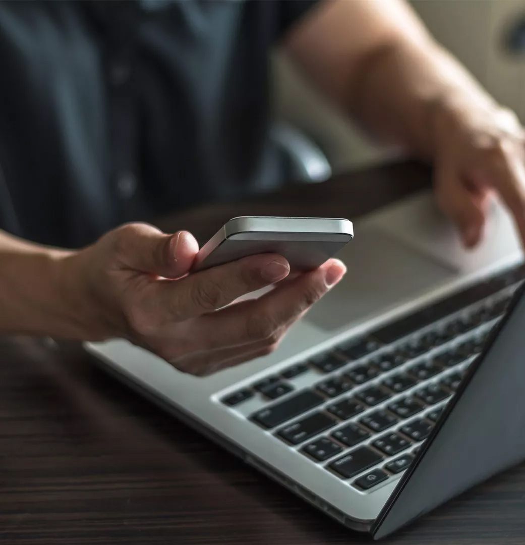 Eine Person hält ein Smartphone in der einen Hand, während die andere Hand mit einem Laptop auf einem Holztisch interagiert.